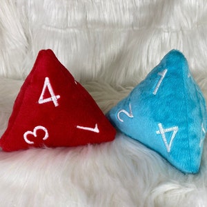 Op de afbeelding: Twee pluche dobbelstenen, een rode en een blauwe, met witte cijfers op elke zijde. De dobbelstenen zijn piramidevormig en liggen op een wit, pluche oppervlak.