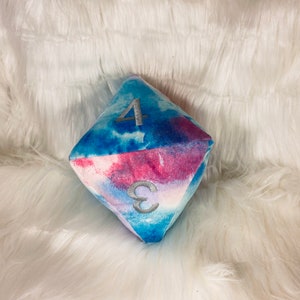 May include: A plush, multi-sided die with a watercolor design in shades of blue and pink. The die features the numbers 3 and 4 in silver embroidery. The soft, geometric shape rests on a fluffy white surface.