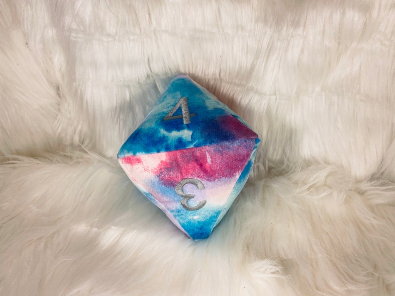 May include: A plush, multi-sided die with a watercolor design in shades of blue and pink. The die features the numbers 3 and 4 in silver embroidery. The soft, geometric shape rests on a fluffy white surface.