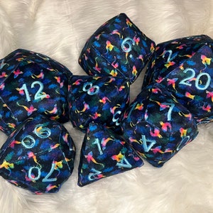 May include: A set of seven plush dice with a dark blue background and a colorful, abstract pattern. The dice are numbered 1, 4, 6, 7, 8, 12, and 20.