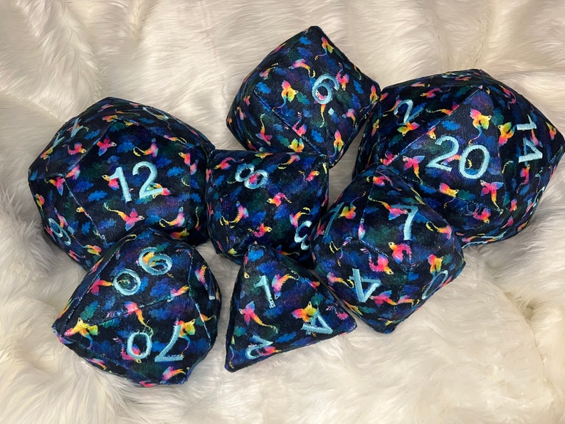 May include: A set of seven plush dice with a dark blue background and a colorful, abstract pattern. The dice are numbered 1, 4, 6, 7, 8, 12, and 20.