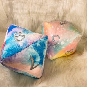 May include: Two plush, multi-sided dice with a watercolor galaxy design. The dice feature embroidered numbers in silver. One die displays the numbers 8 and 1, while the other shows 5 and 10. The dice are soft and colorful.