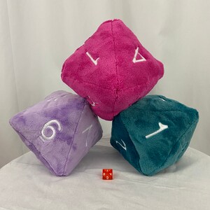 Peut inclure: Trois dés en peluche de couleur rose, violet et turquoise avec des chiffres blancs. Les dés sont disposés en forme de pyramide. Un petit dé en plastique rouge avec des chiffres blancs est au premier plan.