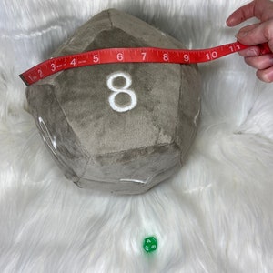 May include: A large, plush, gray d20 die with white embroidered numbers, measuring approximately 10 inches across. A red measuring tape is held across the die. A small green d10 die is also present.