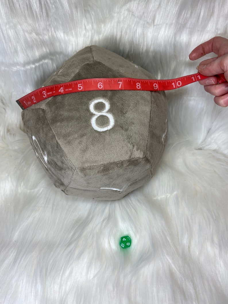 May include: A large, plush, gray d20 die with white embroidered numbers, measuring approximately 10 inches across. A red measuring tape is held across the die. A small green d10 die is also present.