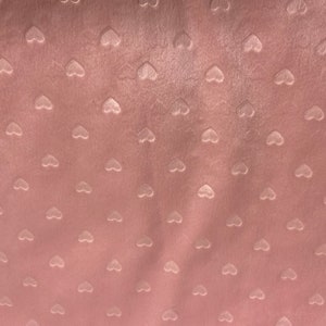May include: Close-up of a soft, pink fabric with a velvety texture. The fabric is patterned with small, raised heart shapes evenly spaced across the surface. The hearts are a lighter shade of pink.