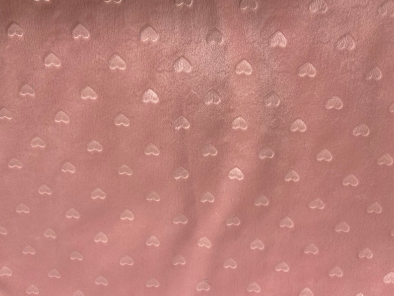 May include: Close-up of a soft, pink fabric with a velvety texture. The fabric is patterned with small, raised heart shapes evenly spaced across the surface. The hearts are a lighter shade of pink.