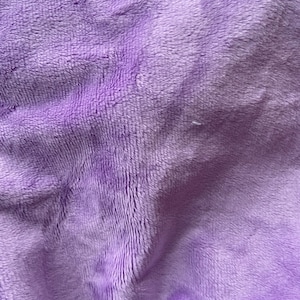 May include: A close-up of a soft, plush fabric in a light purple color. The fabric has a fuzzy texture and is wrinkled, creating a textured appearance.
