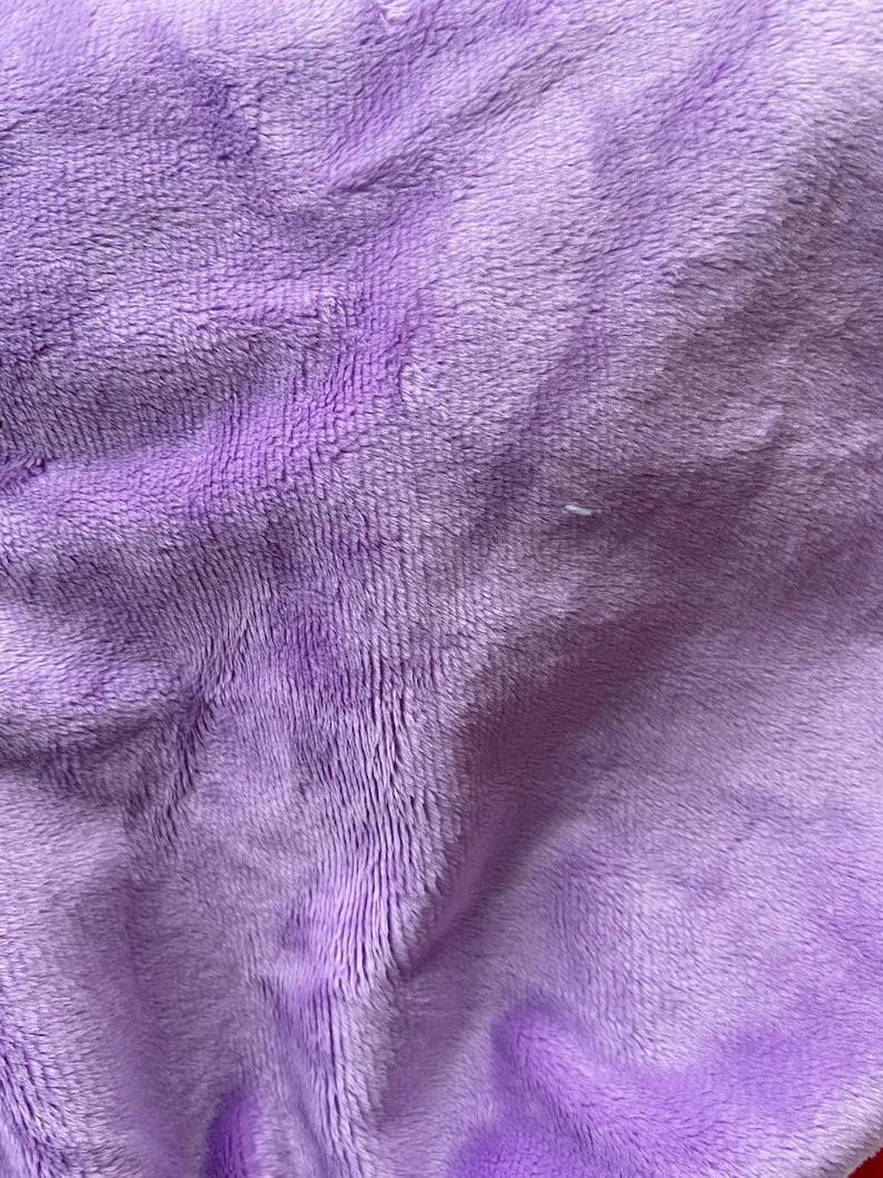 May include: A close-up of a soft, plush fabric in a light purple color. The fabric has a fuzzy texture and is wrinkled, creating a textured appearance.