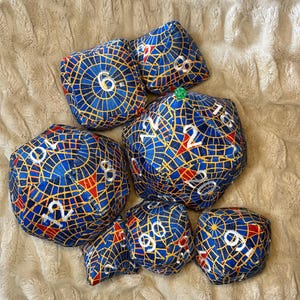 May include: A selection of fabric dice in different sizes, with a blue, yellow, and red mosaic design. The dice display white numbers and are placed on a textured, off-white surface. These dice are suitable for tabletop games.