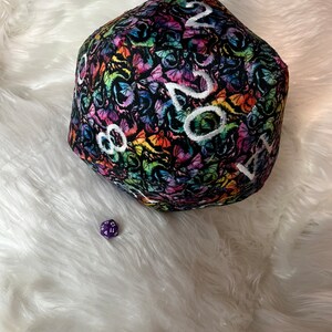 May include: A large, plush, rainbow-colored dice with a black background and white numbers. The dice is covered in a pattern of butterflies. A smaller, purple dice is in the foreground.
