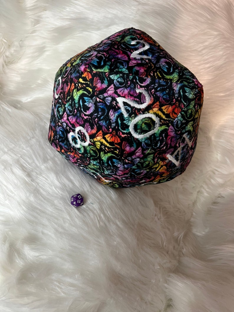 May include: A large, plush, rainbow-colored dice with a black background and white numbers. The dice is covered in a pattern of butterflies. A smaller, purple dice is in the foreground.