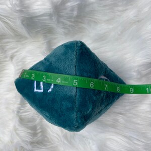 May include: A teal plush die with white embroidered numbers. The die is approximately 4 inches wide. A green measuring tape is held next to the die.