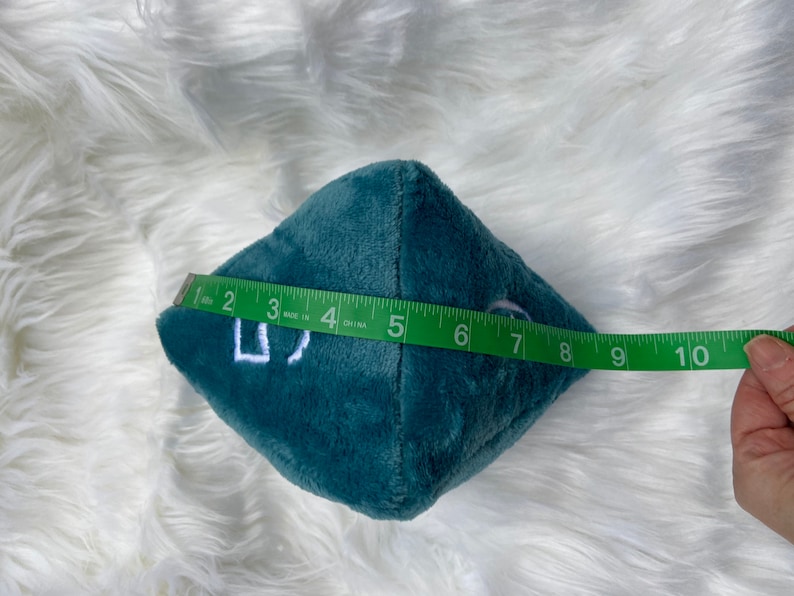 May include: A teal plush die with white embroidered numbers. The die is approximately 4 inches wide. A green measuring tape is held next to the die.