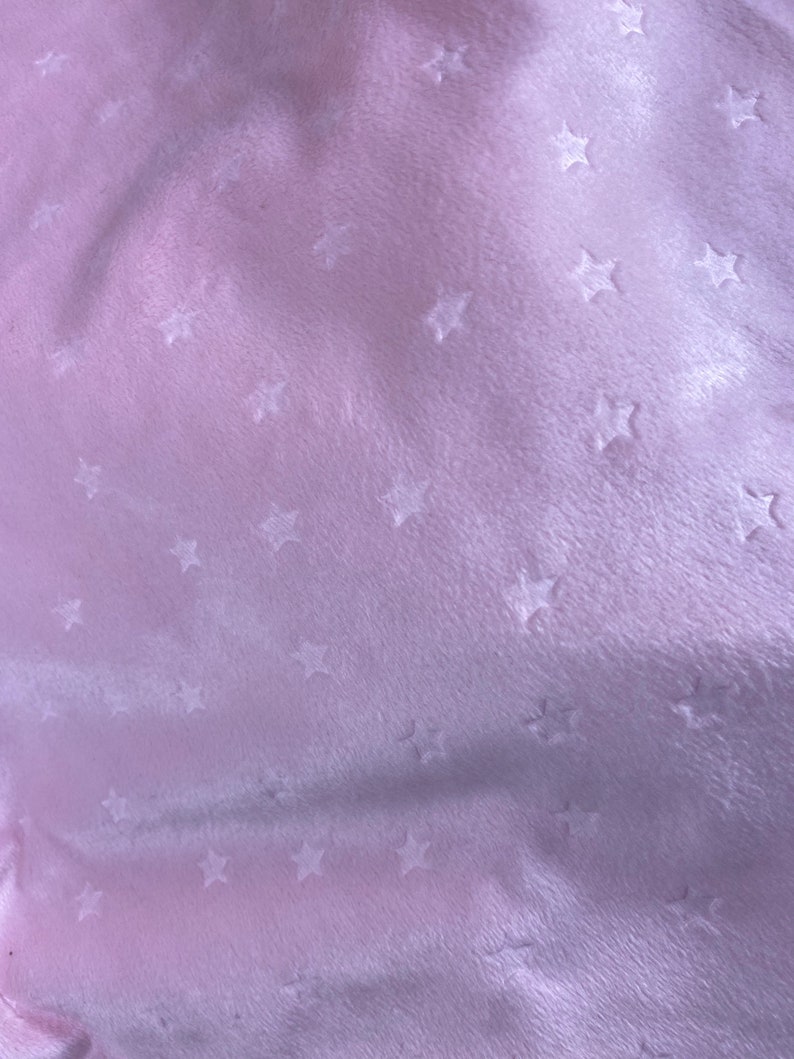 May include: A close-up of a soft, light pink fabric with a velvety texture. The fabric is patterned with small, embossed star shapes scattered across the surface. The lighting creates subtle shadows, enhancing the texture.