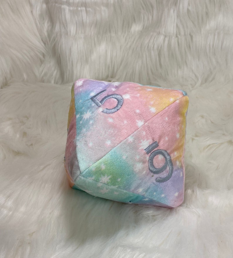 May include: A plush, multi-colored die with embroidered numbers. The die features a pastel rainbow tie-dye pattern with white star accents. The numbers are embroidered in a light gray color.