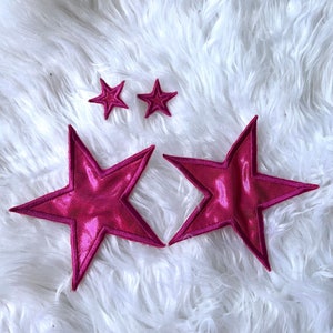 May include: Three pink fabric stars of different sizes. The two smaller stars are on a white fluffy surface. The largest star is in the foreground and is slightly angled.