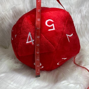 May include: A large, red, plush dice with white numbers 1, 2, 3, 4, 5, and 6. The dice is 7 inches tall.