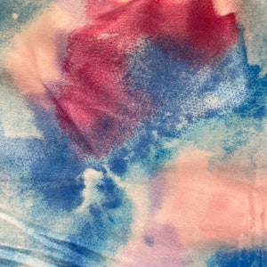 May include: Close-up of a fabric with a watercolor effect. The fabric features a blend of colors, including shades of blue, pink, and purple, creating a soft, blended appearance. The texture appears to be soft and slightly wrinkled, suggesting a comfortable material.