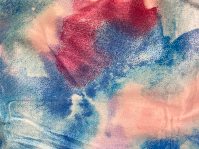 May include: Close-up of a fabric with a watercolor effect. The fabric features a blend of colors, including shades of blue, pink, and purple, creating a soft, blended appearance. The texture appears to be soft and slightly wrinkled, suggesting a comfortable material.