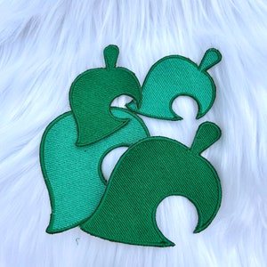 May include: Three green embroidered fabric patches in the shape of a leaf with a curved cut-out. The patches are layered on top of each other.