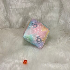 May include: A large, soft, plush D20 die with a pastel galaxy pattern and the numbers 5 and 9. A small, orange, transparent D6 die is also present. Both dice are on a white, fluffy surface.