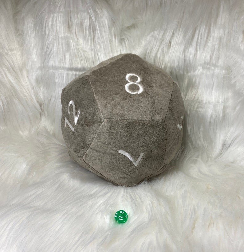 May include: A large, gray plush d12 die with white embroidered numbers. The die is resting on a white, fluffy surface. A small, green d6 die is in the foreground.