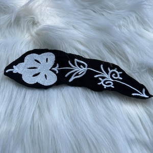 Puede incluir: Un parche de tela negro con bordado blanco de una flor estilizada. La flor tiene una forma redondeada con un diseño de espiral en el centro.
