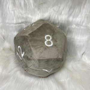 May include: A plush, gray, 12-sided die with white embroidered numbers. The soft, oversized gaming accessory is designed for tabletop role-playing games. The die is approximately 8 inches in diameter.