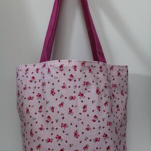 Puede incluir: Un bolso tote rosa con un estampado floral de rosas rojas y peque&ntilde;os detalles morados. El bolso tiene un asa magenta lisa. El bolso es de tela y es adecuado para llevar art&iacute;culos. Dimensiones no visibles.