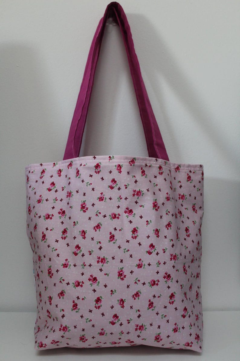 Puede incluir: Un bolso tote rosa con un estampado floral de rosas rojas y peque&ntilde;os detalles morados. El bolso tiene un asa magenta lisa. El bolso es de tela y es adecuado para llevar art&iacute;culos. Dimensiones no visibles.