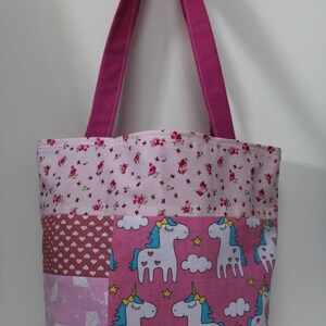 May include: A patchwork tote bag with a pink floral top, a section with hearts, and a pink section with kites and polka dots. The main section features white unicorns with blue manes and yellow horns on a pink background. The bag has magenta handles.