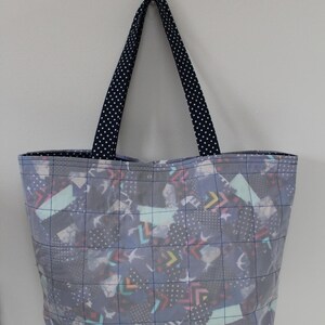 May include: A quilted tote bag with a patchwork design in shades of blue, gray, and white. The bag features a grid pattern and navy blue handles with white polka dots. The bag is designed for carrying items.