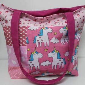 Puede incluir: Un bolso tote rosa con un dise&ntilde;o de patchwork. El bolso presenta un estampado de unicornios sobre un fondo rosa, con crines azules y cuernos amarillos. Las asas son de color magenta liso y el bolso est&aacute; forrado con un estampado floral.