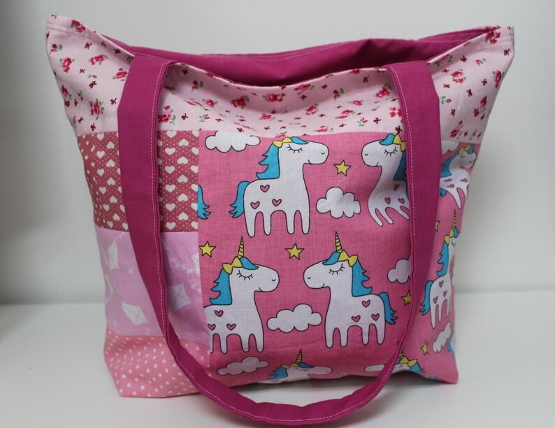 Puede incluir: Un bolso tote rosa con un dise&ntilde;o de patchwork. El bolso presenta un estampado de unicornios sobre un fondo rosa, con crines azules y cuernos amarillos. Las asas son de color magenta liso y el bolso est&aacute; forrado con un estampado floral.