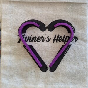 May include: A purple twine helper tool in the shape of a heart. The tool is on a white canvas bag with the text "Twiner's Helper" printed on it.