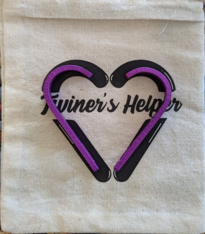 May include: A purple twine helper tool in the shape of a heart. The tool is on a white canvas bag with the text "Twiner's Helper" printed on it.