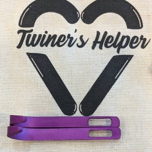 May include: Two purple metal tools with a rounded end and a rectangular hole. The tools are on a white canvas bag with the text "Twiner's Helper" in black.