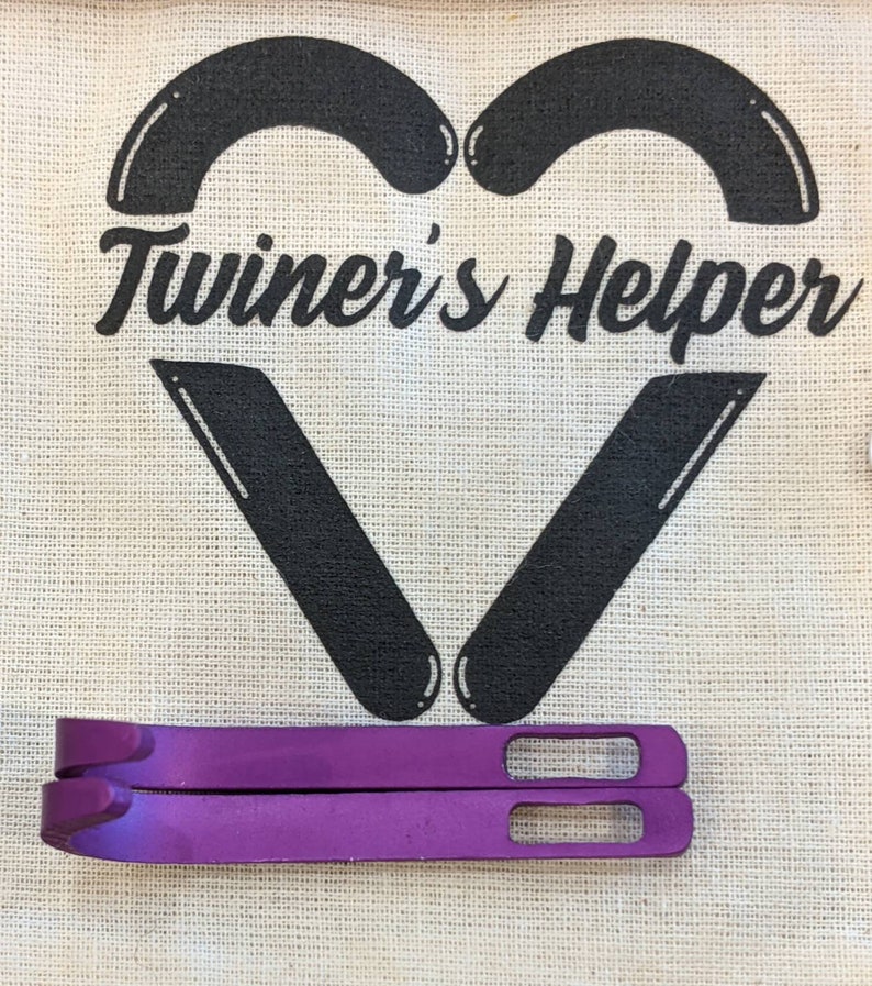 May include: Two purple metal tools with a rounded end and a rectangular hole. The tools are on a white canvas bag with the text "Twiner's Helper" in black.