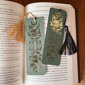 May include: Two decorative bookmarks with gold foil designs of deer, butterflies, and architectural elements. The bookmarks are a muted teal color and have matching tassels. One bookmark has a tan tassel, and the other has a black tassel.