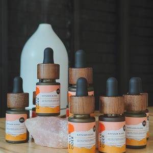 Può includere: Una collezione di piccole bottiglie di vetro marrone con tappi di legno e contagocce neri, etichettate "Mulled Cider Diffuser Blend". Sono disposte su una superficie di legno con un diffusore bianco e un cristallo rosa. Le etichette hanno accenti arancioni e bianchi.