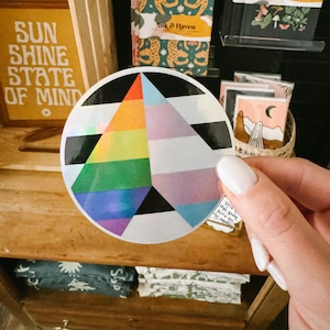 May include: A round sticker with a rainbow and transgender pride flag design. The sticker has a black and white striped background.