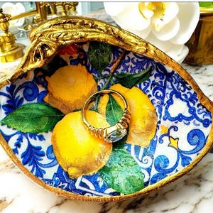 Op de afbeelding: Een decoratieve schelpenringschotel met een gouden rand. De binnenkant heeft een citroen- en bladerenontwerp op een blauw-witte achtergrond. Een gouden ring met een blauwe edelsteen ligt erin.