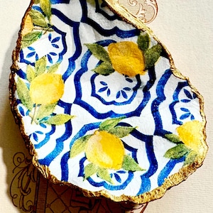 Può includere: Un piatto in ceramica bianca a forma di conchiglia di ostrica con un motivo geometrico blu e bianco e limoni gialli dipinti su di esso. La conchiglia è rifinita con vernice dorata.