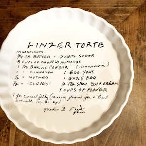 May include: A white pie plate with a handwritten recipe for Linzer Torte. The recipe includes ingredients like butter, sugar, almonds, baking powder, cinnamon, nutmeg, cloves, eggs, sour cream, and flour. It also includes instructions for making a currant jelly filling.