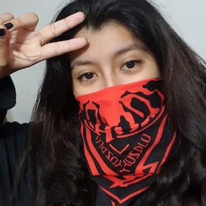 Può includere: Una bandana rossa e nera con un design grafico copre la parte inferiore del viso. La bandana ha un motivo e un testo ripetuti. La persona indossa un top nero e ha lunghi capelli scuri. La persona sta facendo un segno di pace.