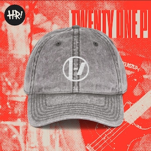Könnte beinhalten: Graue Baseballkappe mit weißem Logo und dem Text "TWENTY ONE P!" auf rotem Hintergrund. Die Kappe hat einen gebogenen Schirm und ein kreisförmiges Logo. Der Hintergrund hat ein strukturiertes Design mit den Buchstaben "HR!" in einem schwarzen Kreis.