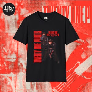 May include: Black t-shirt with a red and white graphic design. The shirt features the text "Twenty One Pilots" in red vertical lettering. The image on the shirt shows two band members and the text "The Clancy Tour Breach 2023".