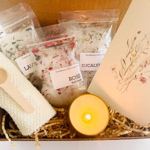 May include: A gift box filled with bath salts in various scents, including lavender, rose, and eucalyptus. The box also includes a small wooden spoon, a white washcloth, and a candle.
