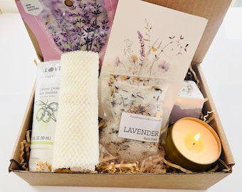 May include: A gift box filled with lavender-themed bath and body products. The box includes a lavender bath soak, a hand cream, a loofah, and a candle. The box is decorated with a lavender-coloured ribbon.
